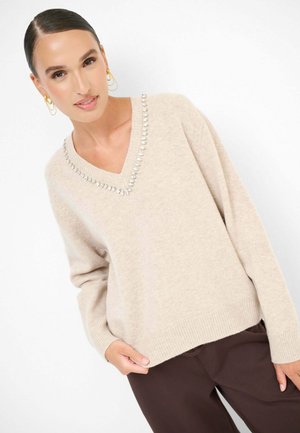 Maglione beige lavorato a maglia con scollatura a V, impreziosito da borchie metalliche lungo il colletto. Maniche lunghe e orlo e polsini a costine.