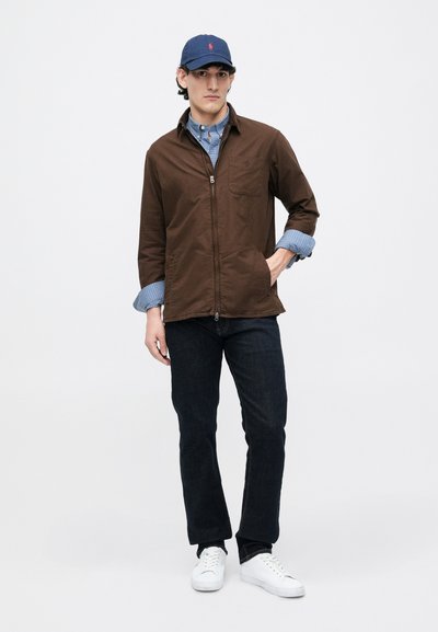 Jaqueta castanha com fecho zip, com um bolso no peito, sobre uma camisa azul xadrez, combinada com jeans escuros e ténis brancos, complementada com um boné azul-marinho.