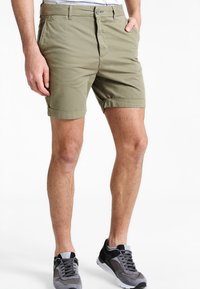 Shorts vert olive en tissu léger, dotés d'une fermeture à bouton, de ourlets roulés et de poches latérales, associés à des chaussures de sport grises.