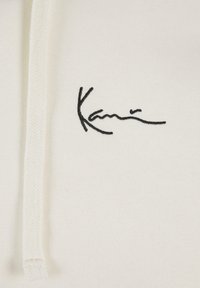 Hvit hettegenser med svart brodert signatur. Materialet ser mykt ut, med et enkelt design og snøringdetaljer.