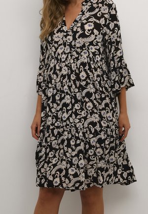 Kobieta nosząca czarną sukienkę do kolan z kremowymi i niebieskimi wzorami kwiatowymi oraz rękawami o długości trzech kwartalnych, stojąca na tle białym.