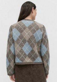 Kvinde iført en blå og brun argyle-mønstret sweater, set fra ryggen mod en ensfarvet hvid baggrund.