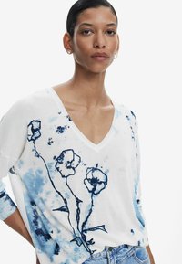 Weißer Pullover mit tiefem V-Ausschnitt und langen Ärmeln, versehen mit blauen Blumenmustern und Aquarellakzenten. Texturiertes Material mit lockerer Passform.