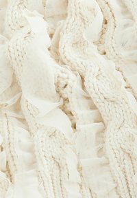 Texturerad krämfärgad tyg med stickade mönster och mjuka volangdetaljer i en flödande arrangemang. Lämplig för heminredning eller modeartiklar.