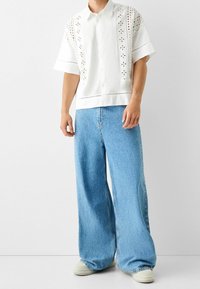 Bílá košile s krátkým rukávem s výšivkou očkování a zapínáním na knoflíky, v kombinaci s volnými světlými modrými džínami a béžovými teniskami.