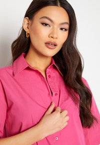 Camicia rosa, con bottoni e colletto a punta, a maniche corte; tessuto morbido e design aderente. Grandi orecchini a cerchio dorati completano il look.