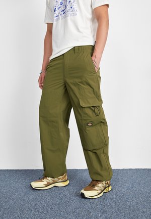 Osoba nosząca oliwkowe spodnie typu cargo z wieloma kieszeniami, beżowo-brązowe buty sportowe oraz białą koszulkę New Balance z grafiką.