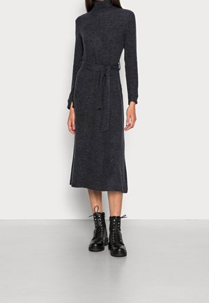 Kobieta w ciemnoszarej sukience midi z długim rękawem, z pasującym paskiem oraz czarnych sznurowanych botkach, stoi na jasnoszarej podłodze.