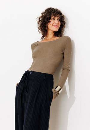 Femme aux cheveux bouclés s'appuyant contre un mur blanc, portant un haut marron décolleté, un pantalon large à rayures noires et un bracelet en or.