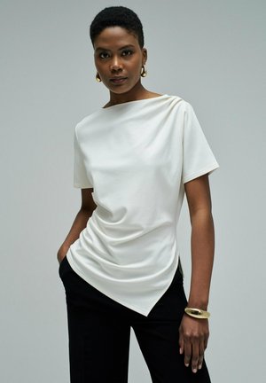 Mujer con camiseta blanca asimétrica de manga corta, pantalones negros, pendientes de aro dorados y una pulsera dorada, posando frente a un fondo liso.