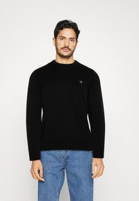 Suéter negro de manga larga con cuello redondo y un pequeño logo bordado en el pecho, combinado con unos jeans de denim azul claro.