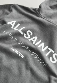 Sweat à capuche gris avec "ALLSAINTS" en grandes lettres blanches, des caractères japonais en dessous, et "LONDON" en plus petit texte blanc en dessous.