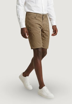 Hombre caminando, vestido con pantalones cortos marrones hasta la rodilla, camisa blanca de manga larga y zapatillas blancas sobre un fondo liso.