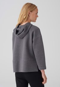 Felpa grigia con un tessuto testurizzato, dotata di cappuccio posteriore, spalle scese e spacchi laterali all'orlo. Le maniche sono leggermente arrotolate.