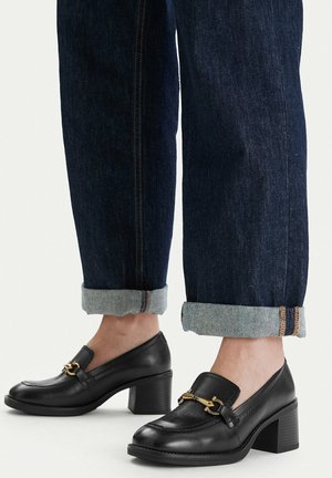 Schwarze Loafers aus Leder mit Blockabsatz und goldener Kettendetail, getragen mit aufgekrempelten dunkelblauen Jeans an nackten Füßen.