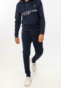 Marineblaues Sweatshirt und -hosen-Set mit Logo der U.S. Polo Assn., das eine Kapuze und seitliche Streifen aufweist. Kombiniert mit weißen Sneakers.