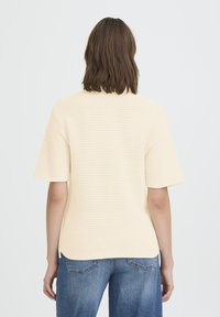 Haut en maille beige clair à manches courtes avec un motif texturé. Présente une coupe ample et un ourlet légèrement arrondi à l'arrière. Porté avec un jean bleu.