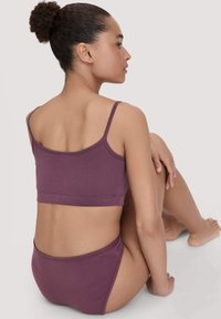 Vrouw met krullend haar in een bun, zittend en gekleed in een bijpassende paarse crop top en briefjes tegen een effen lichte achtergrond.