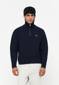 Navyblauwe gebreide trui met hoge ritskraag, gekenmerkt door een textuurpatroon en een klein groen logoaccent op de borst.