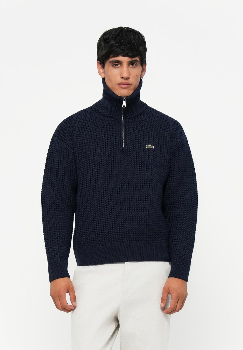 Navyblauwe gebreide trui met hoge ritskraag, gekenmerkt door een textuurpatroon en een klein groen logoaccent op de borst.