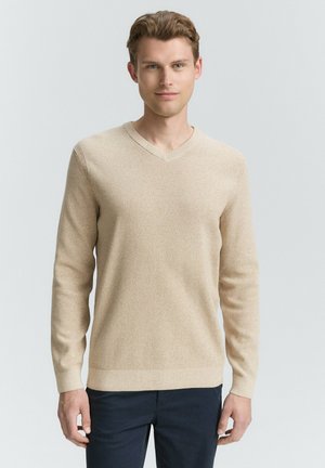 Helles beige V-Ausschnitt-Pullover mit langen Ärmeln, strukturiertem Strickmuster und gerippten Bündchen. Über dunkelblauen Hosen getragen vor einem neutralen Hintergrund.