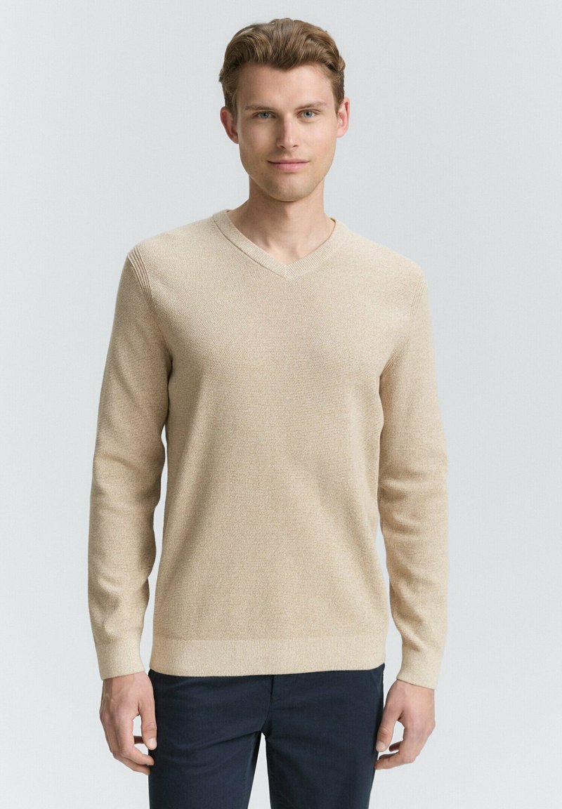 Helles beige V-Ausschnitt-Pullover mit langen Ärmeln, strukturiertem Strickmuster und gerippten Bündchen. Über dunkelblauen Hosen getragen vor einem neutralen Hintergrund.