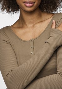 Langermet ribbestrikket topp i dempet brun med scoop hals og fem dekorative knapper nedover i midten. Myk og elastisk stoffstruktur.