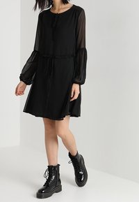 Schwarzes Kleid mit durchsichtigen, puffigen Ärmeln, Rüschendetails, einer Bindung am Kragen und einem fließenden Rock, kombiniert mit schwarzen Schnürstiefeletten.