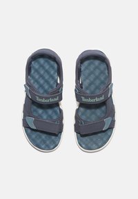 Modré a šedé sandály s nastavitelnými popruhy, texturovanou stélkou a bílou podrážkou; název značky "Timberland" umístěn na popruzích.