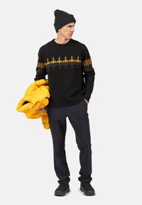 Czarny sweter z żółto-szarym wzorem falistym, w połączeniu z granatowymi spodniami, szarym beanie i trzymający złożoną żółtą kurtkę puchową.