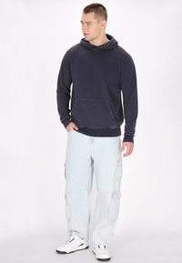 Felpa blu navy con tasca anteriore, tessuto texturizzato, abbinata a pantaloni cargo azzurri chiari con numerose tasche e vestibilità ampia. Sneakers bianche casual.