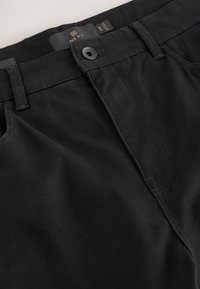 Gros plan de la ceinture d'un jean en denim noir avec bouton fermé, passants de ceinture et étiquette de taille visible à l'intérieur.
