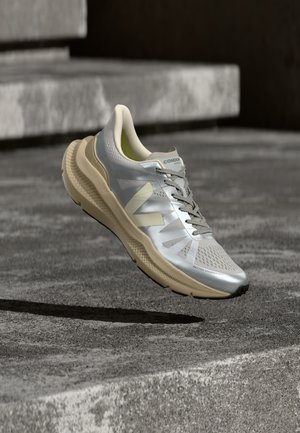 Hõbedase ja beeži jooksujalats tekstuurse tallaga, paelte ja brändinguga, hõljumas kareda betoonpinna ja astmete kohal.