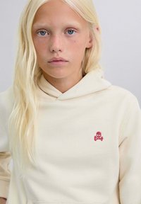 Joven con cabello rubio y ojos azules, vistiendo una sudadera crema con un pequeño bordado de una calavera y huesos cruzados en el pecho.