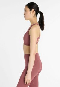 Nuori nainen, jolla on musta ponihäntä, seisoo profiilissa pukeutuneena mauven väriseen urheiluliiviin ja vastaaviin korkeavyötäröisiin legginsseihin, taustalla on pelkkä valkoinen tausta.