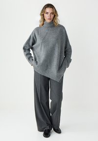 Jasnoszary, oversize sweter z wysokim kołnierzem i asymetrycznym dołem, w połączeniu z ciemnoszarymi spodniami w szerokiej nogawce i czarnymi butami.