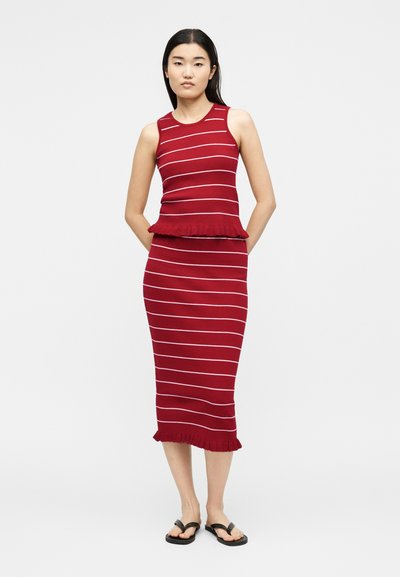 Γυναίκα όρθια με τα χέρια πίσω από την πλάτη, φοράει ένα αμάνικο κόκκινο φόρεμα με λευκές οριζόντιες ρίγες και βολάν στο τελείωμα, μαύρα σανδάλια.