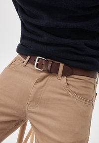 Bež hlače iz bombaža imajo sproščen kroj z sprednjimi žepi. Temno rjavi usnjen pas je zavarovan s srebrno kovinsko sponko.