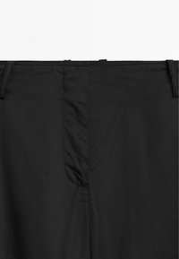 Gros plan sur la ceinture d’un pantalon noir avec passants pour ceinture et une fermeture éclair dissimulée à l’avant.