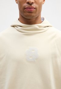 Beżowa bluza z kapturem o miękkiej teksturze i luźnym kapturze. Z przodu znajduje się jasnoszary, puszysty logo "8".