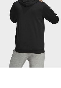 Persona che indossa un hoodie nero con strisce bianche sulle maniche e pantaloni grigi, voltato di spalle con le mani vicino alla testa.