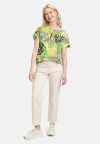 Kolorowa krótka koszulka w zielono-żółte wzory z motywem liści, połączona z jasnobeżowymi spodniami o prostym kroju i jasnymi trampkami.