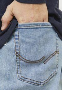 Jeans de mezclilla azul claros con un bolsillo trasero que presenta costuras naranjas. Mano parcialmente visible, agarrando el dobladillo de una blusa oscura.
