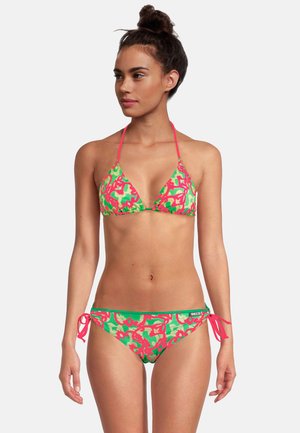 Femme portant un bikini à imprimé floral vert et rose avec des bretelles à nouer, debout devant un fond clair uni, regardant sur le côté.