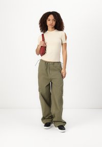 Beige rilletta t-skjorte med korte ermer og utklipte skuldre, olivengrønne vide cargo-bukser og svarte joggesko. Modellen holder en rød veske.