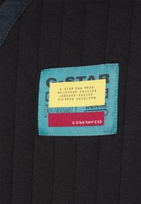 G-Star Raw tygmärke på en svart, ribbad textil. Märket har en teal bakgrund med röd accent, och innehåller varumärkes- och skötselinformation.