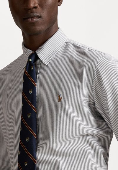 Kreklains ar pelēkām un baltām vertikālām svītrām, uz krūtīm ir neliela izšūta logo, kā arī zilā kaklasaite ar rakstainiem akcentiem.