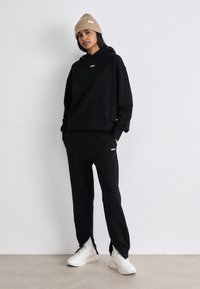 Melnas kapuces un sporta bikses komplekts, kurā ir zelta "BOSS" logo akcenti. Modelis valkā beidzīgu adītu cepuri un baltus sneakers. Gluda auduma tekstūra.