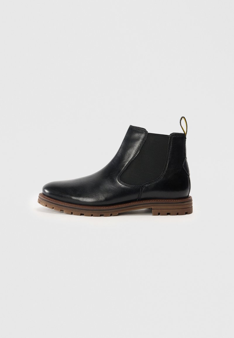 Botín Chelsea de cuero negro con paneles laterales elásticos, punta redonda y suela de goma marrón texturizada. Presenta una lengüeta amarilla en el talón.