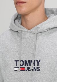 Sudadera gris con cordones. Presenta el texto "TOMMY JEANS" impreso en azul marino con un acento de logo en rojo y blanco. Tela suave y texturizada.